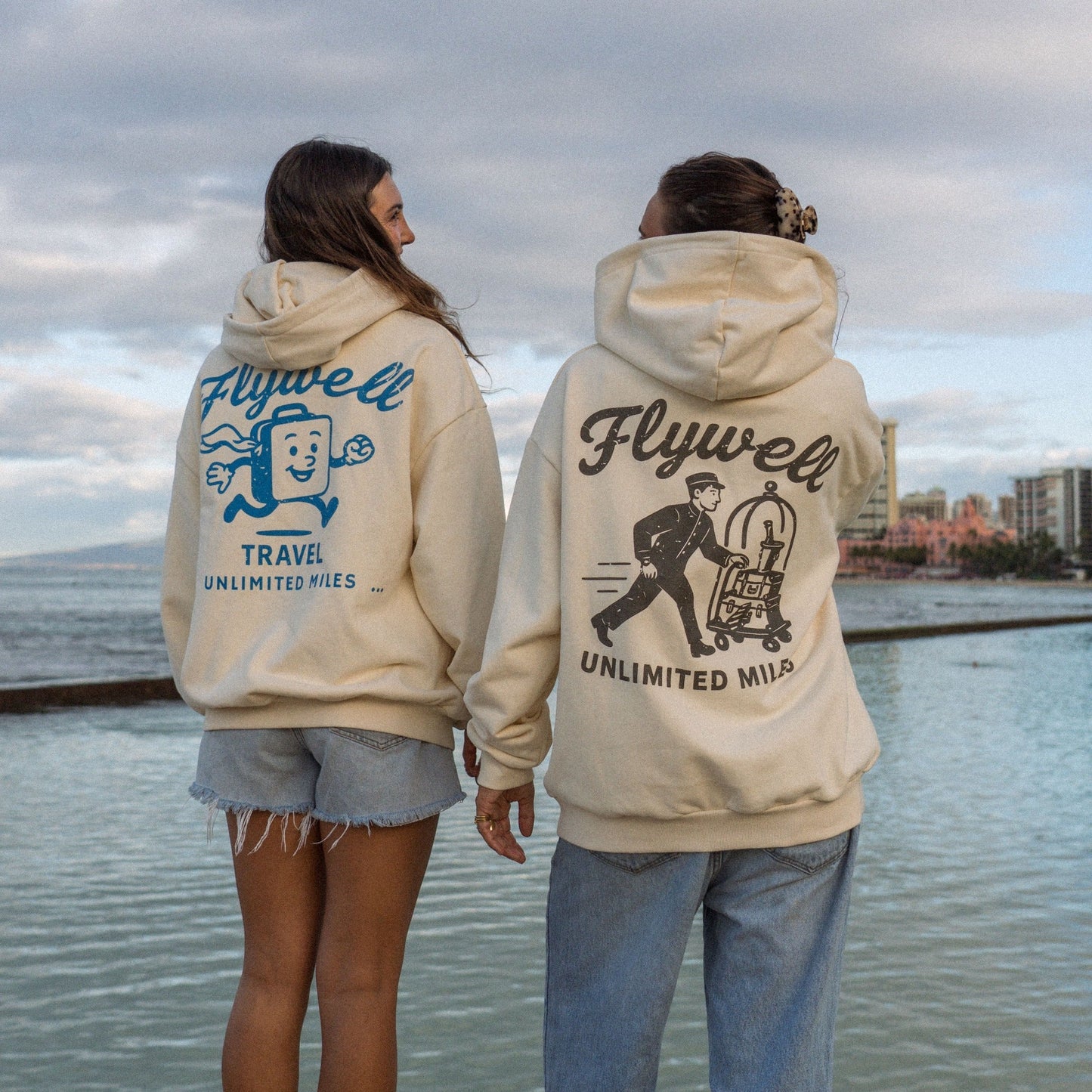 Two people wearing Flywell hoodies by a waterfront with a cloudy sky.