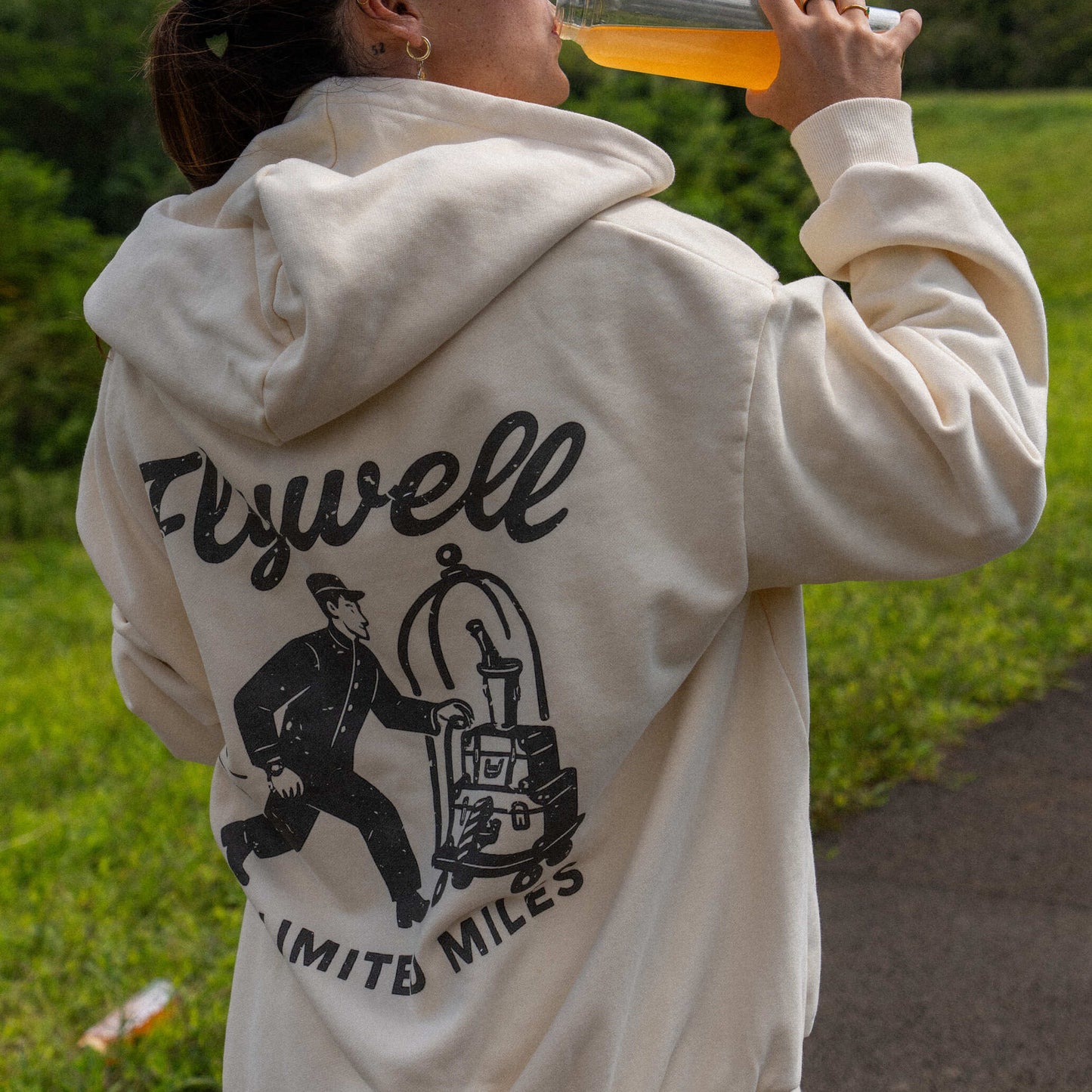 Person wearing a hoodie with 'Flywell' branding, drinking from a bottle outdoors.