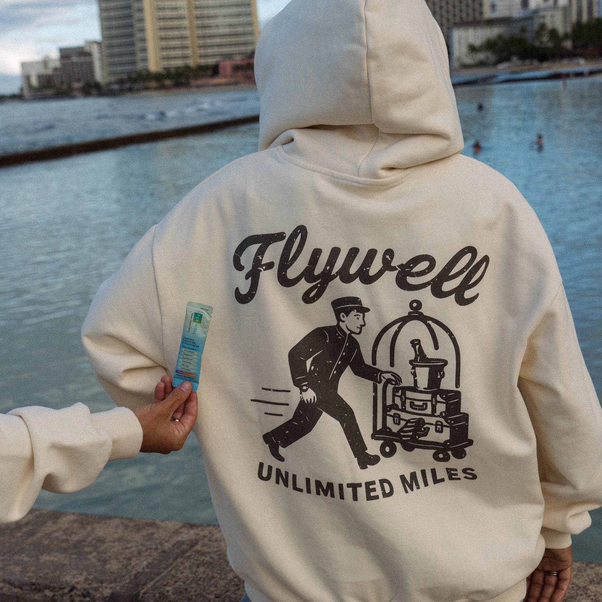 Person wearing a hoodie with 'Flywell Unlimited Miles' design by a waterfront.